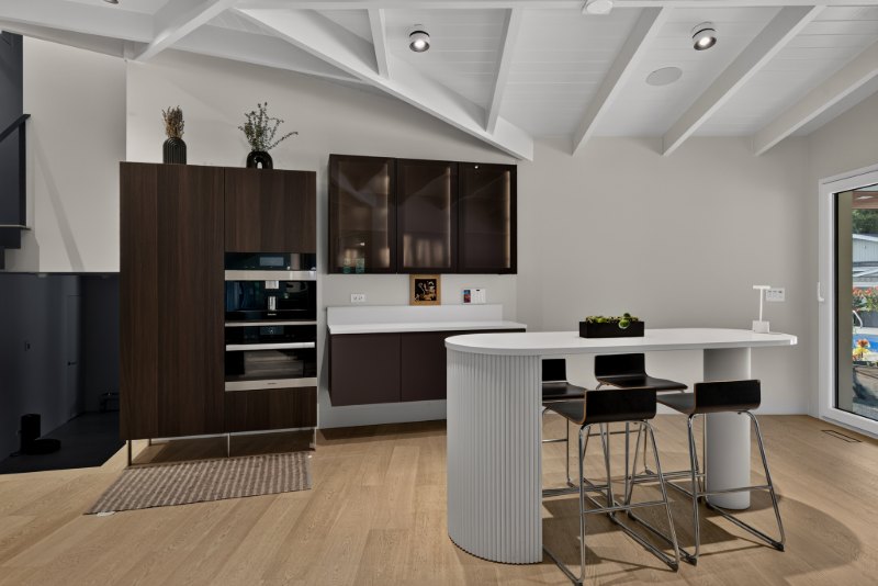 Bronze glass display cabinets above a floating counter, beside tall dark wood cabinetry with a coffee machine and ovens, and an off-white table with a fluted end and a round leg.