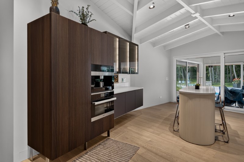 Tall dark wood cabinetry with a built-in coffee machine and ovens, beside metal-framed bronze glass cabinets lit from within, and an off-white fluted table.