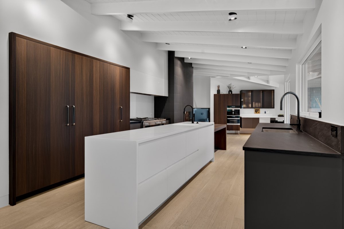 Kitchen with tall fumed oak refrigerator cabinetry, a white waterfall island and dark graphite cabinetry along the sink wall, with bronze glass display cabinets at the far end under a white beamed ceiling.
