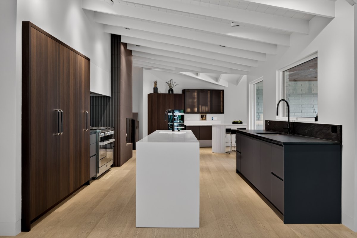 View down the full length of the kitchen: fumed oak tall cabinetry to the left, a white island in the center, dark graphite cabinetry to the right, and bronze glass cabinets and an off-white fluted table beyond.