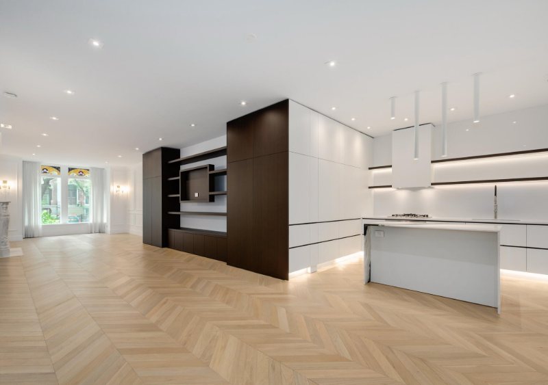 Open-plan white kitchen with a Wenge veneer media and shelving volume, across chevron wood flooring.
