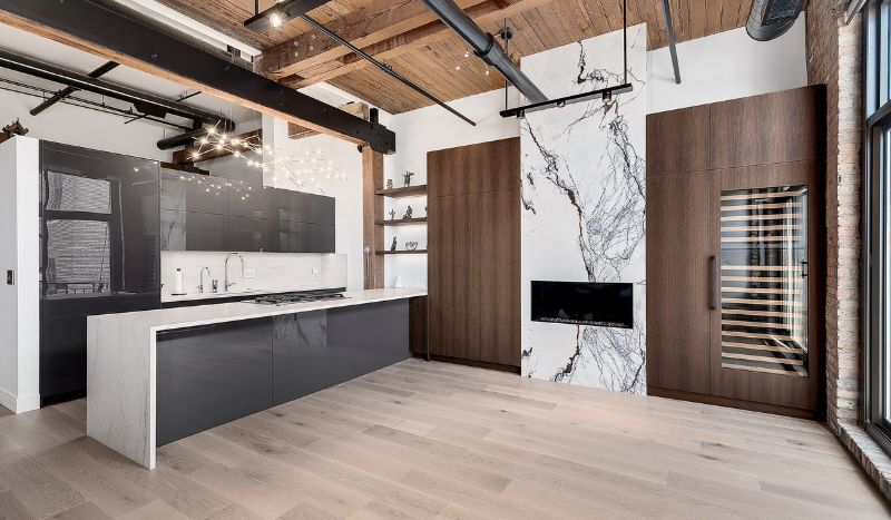 Loft kitchen with dark gloss cabinetry, a white stone waterfall island and dark wood millwork beside a book-matched stone feature wall, under exposed timber beams.